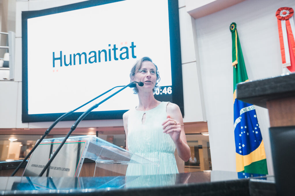 A arquiteta Carolina Nunes aparece em 1o plano discursando na tribuna da ALESC. Ao fundo, aparece o slide no telão, onde está escrito "Humanitat". A bandeira do Brasil também aparece atrás da tribuna.