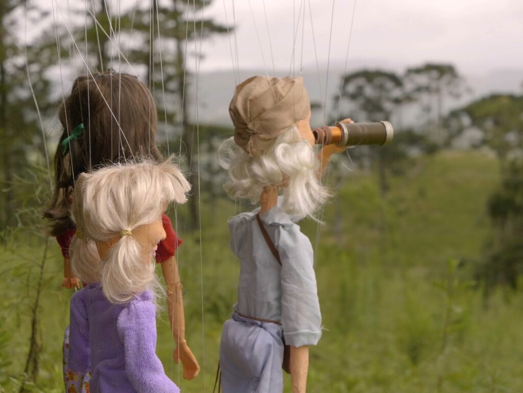 3 bonecos de marionetes estão de costas, observando uma paisagem de araucárias. Um dos bonecos, representando Fritz Müller, observa com luneta.