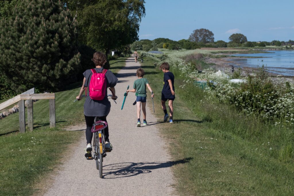 A imagem mostra um caminho rústico, porém muito confortável para pedestres e ciclistas, e à direita do caminho há uma frente de água, que pode ser uma baía ou uma lagoa. No caminho, há em primeiro plano uma ciclista com mochila rosa. Um pouco na frente, há duas crianças com brinquedos, e mais na frente, há uma mulher passeando com um cachorro. A transição entre o caminho e a lagoa ou baía é uma área natural com vegetação baixa. O céu está azul.